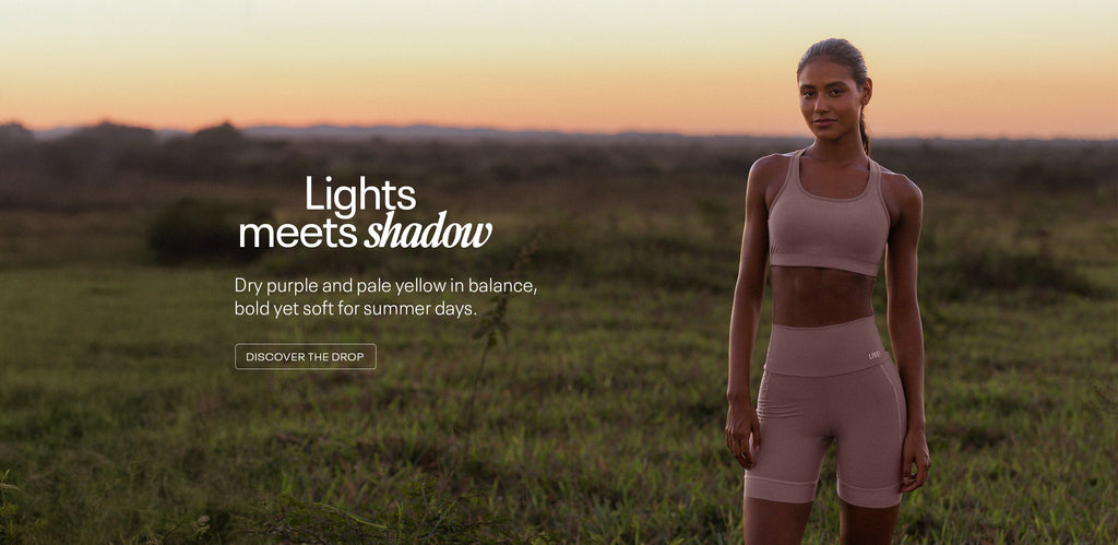 A woman stands confidently in a field at sunset, wearing a matching dry purple sports bra set from LIVE! Activewear. The warm, natural lighting and soft horizon reflect the collection’s theme, “Lights meets shadow,” blending elegance and strength in a serene outdoor setting.