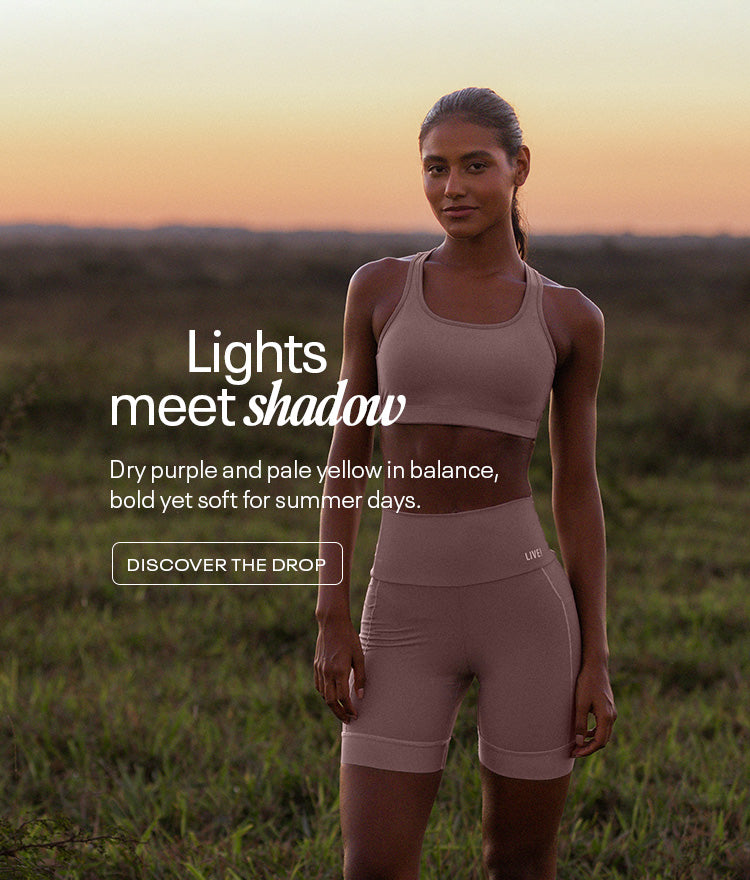 A woman stands confidently in a field at sunset, wearing a matching dry purple sports bra set from LIVE! Activewear. The warm, natural lighting and soft horizon reflect the collection’s theme, “Lights meets shadow,” blending elegance and strength in a serene outdoor setting.