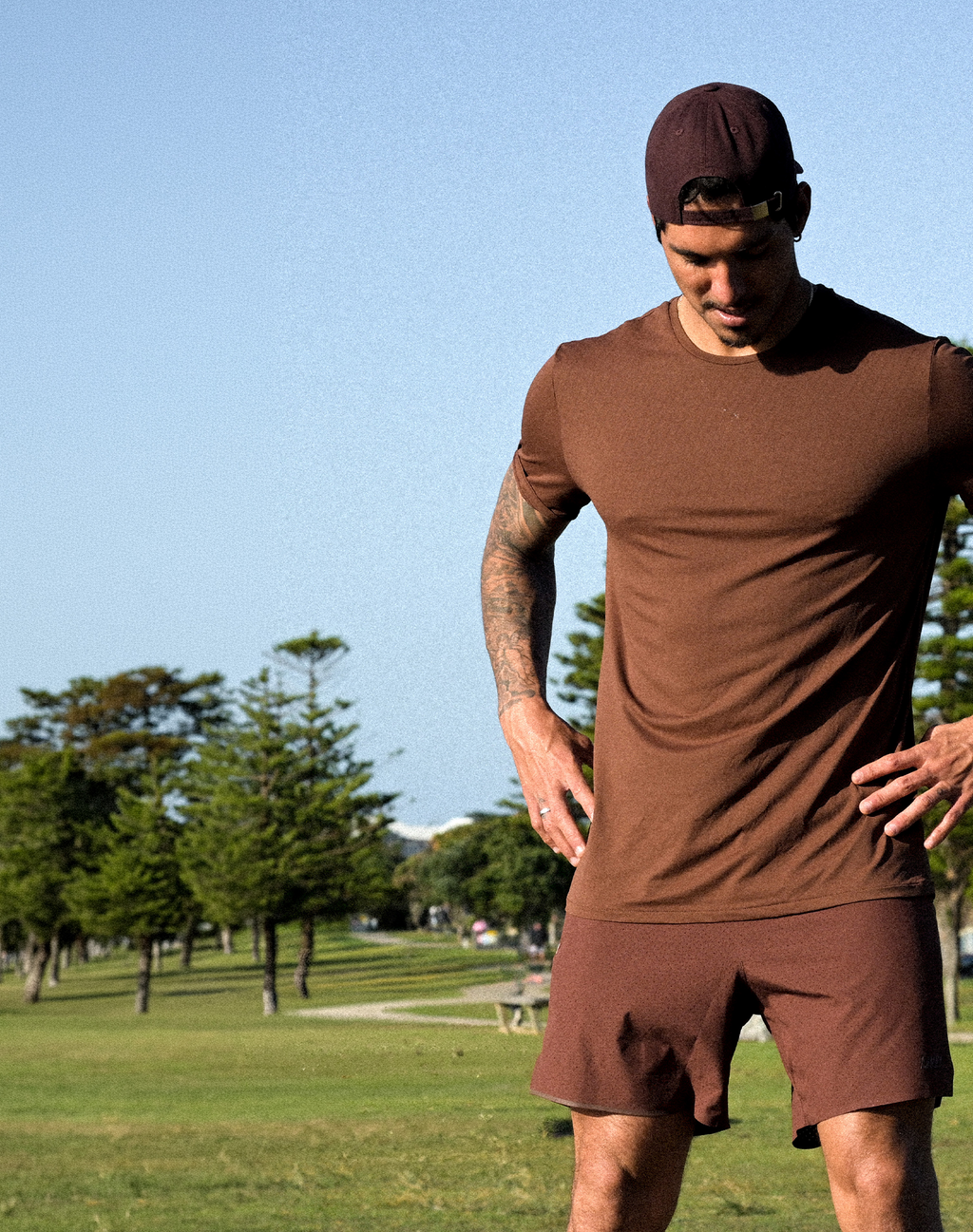 A man wearing a matching brown athletic t-shirt and shorts set, standing outdoors in a park with trees and a blue sky in the background.