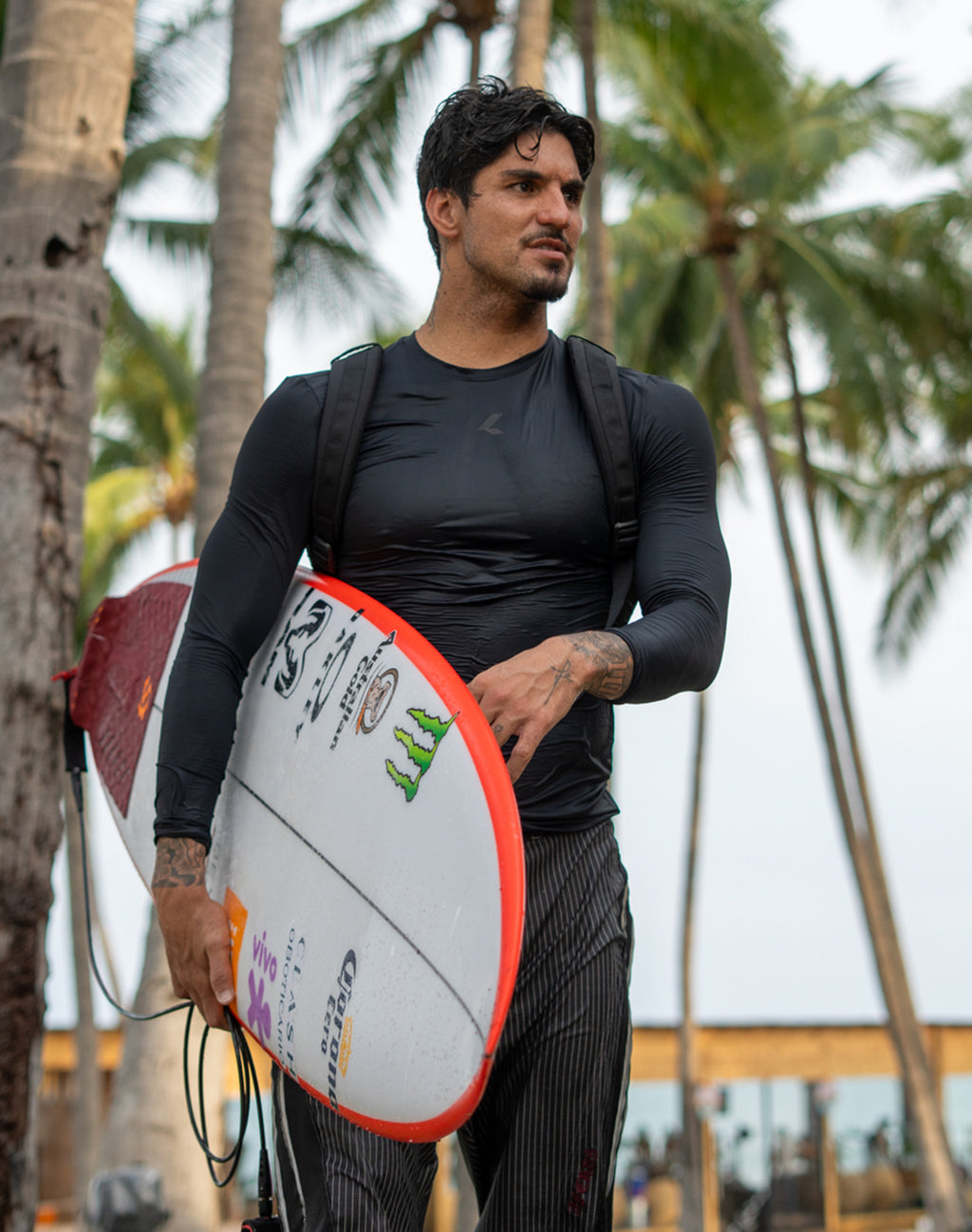 Man wearing a black performance top carrying a surfboard on a palm-lined beach.