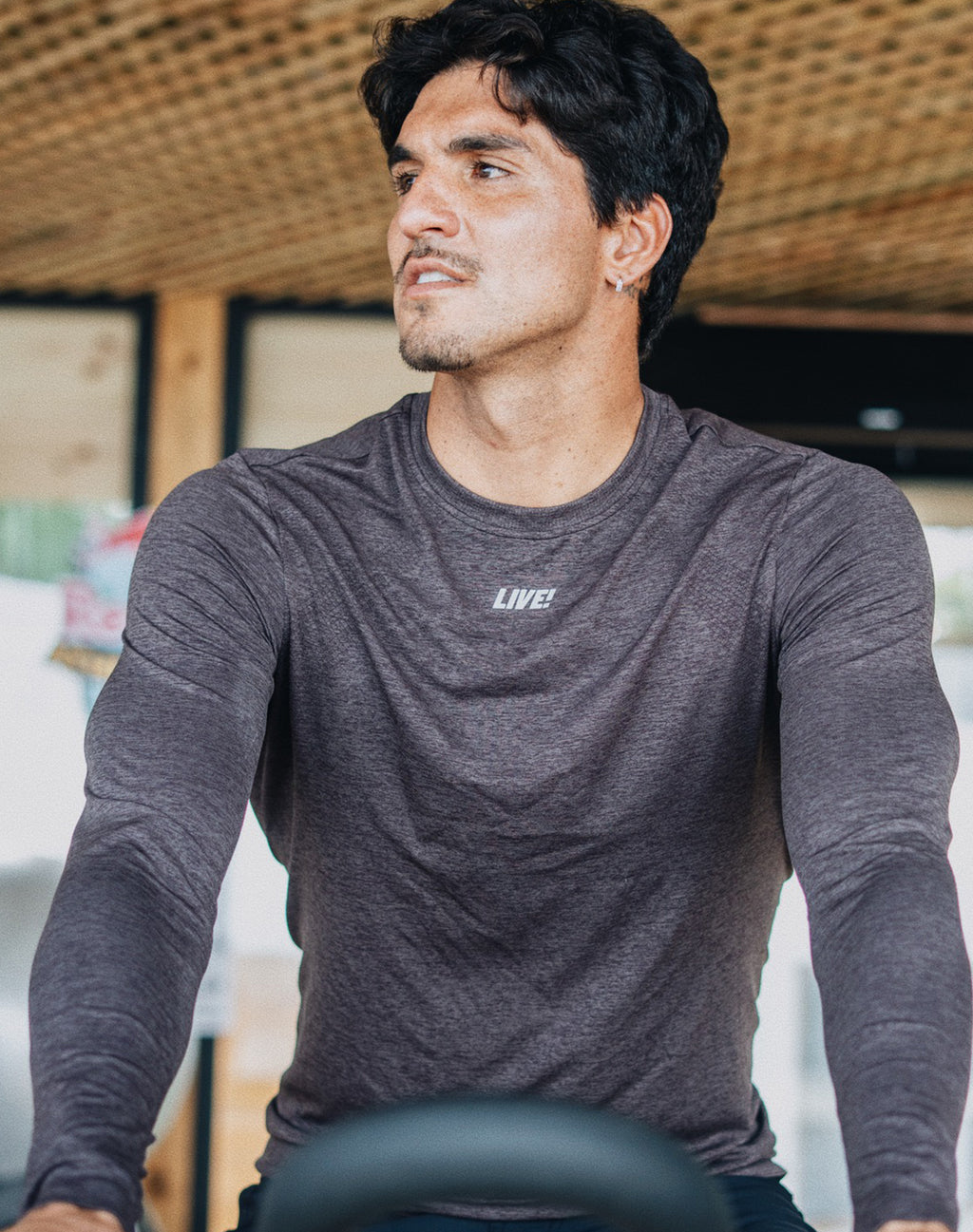 A man wearing a dark gray heathered long-sleeve athletic shirt with a small white 'LIVE!' logo on the chest, riding a stationary in a gym setting.
