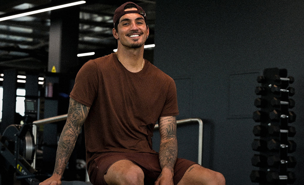A smiling man with tattoos sits on a gym bench wearing a brown t-shirt, matching shorts, and a dark cap in a gym setting with dumbbells visible in the background.