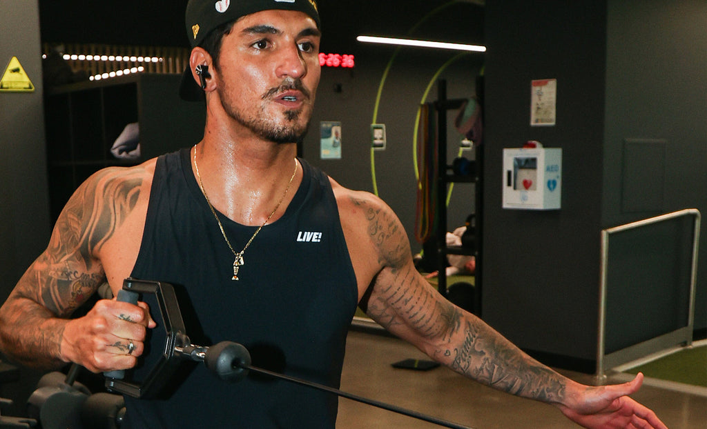 A man with tattoos wearing a black sleeveless tank top with a white 'LIVE!' logo, a black cap, and a necklace, exercising with a cable machine in a gym.