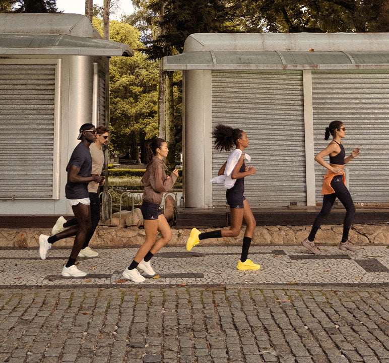 Five runners jogging side by side on a cobblestone street in front of modern metal shuttered buildings with trees in the background.