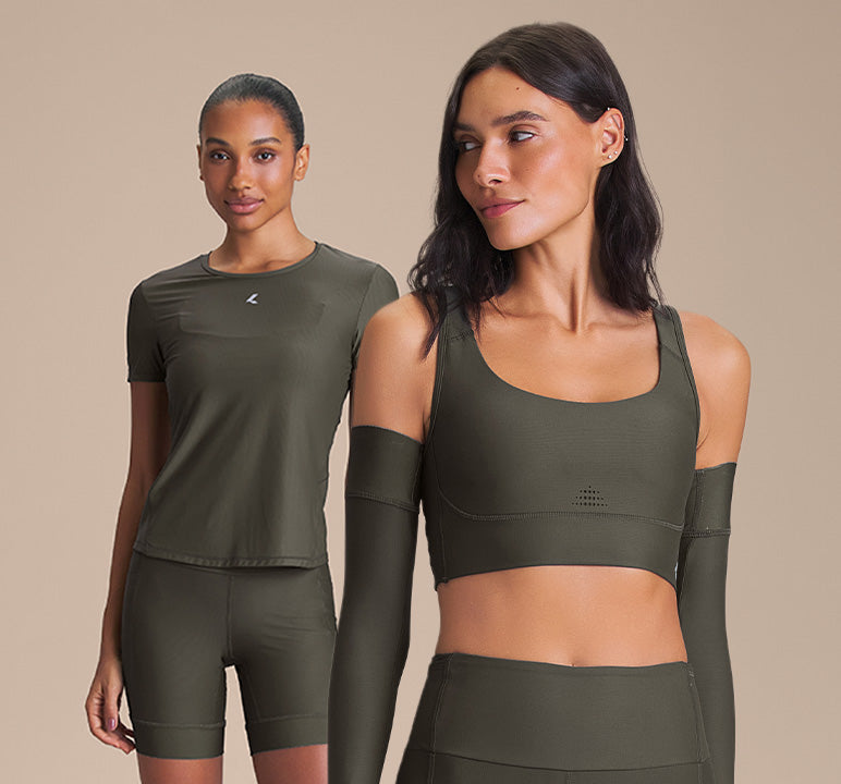 Two women modeling matching olive green athletic wear against a plain beige studio background. One wears a short-sleeve crop top and biker shorts, and the other wears a cold-shoulder crop top with matching arm sleeves and high-waisted bottoms.
