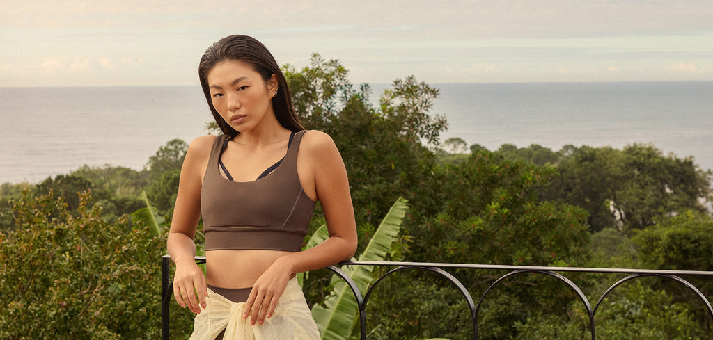 A woman wearing a dark brown sports bra and a cream skirt stands outdoors on a balcony overlooking a lush green landscape and the ocean.