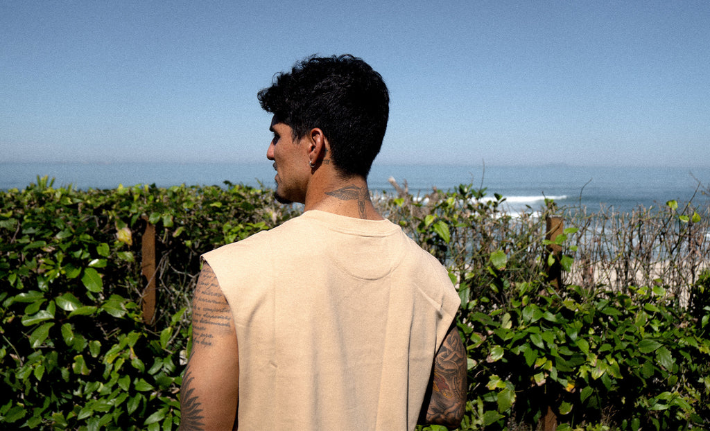 A man with tattoos is seen from behind, wearing a beige sleeveless t-shirt, standing outdoors in front of green bushes with a beach and ocean in the background under a clear blue sky.