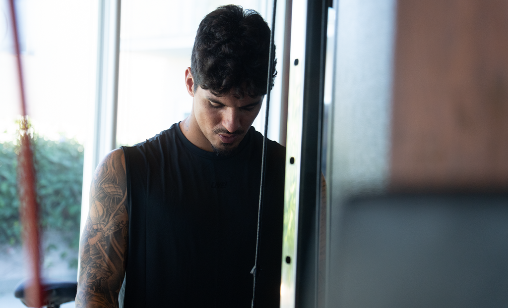 A man with a tattooed arm wearing a black sleeveless shirt, looking down while using a cable machine in a gym.