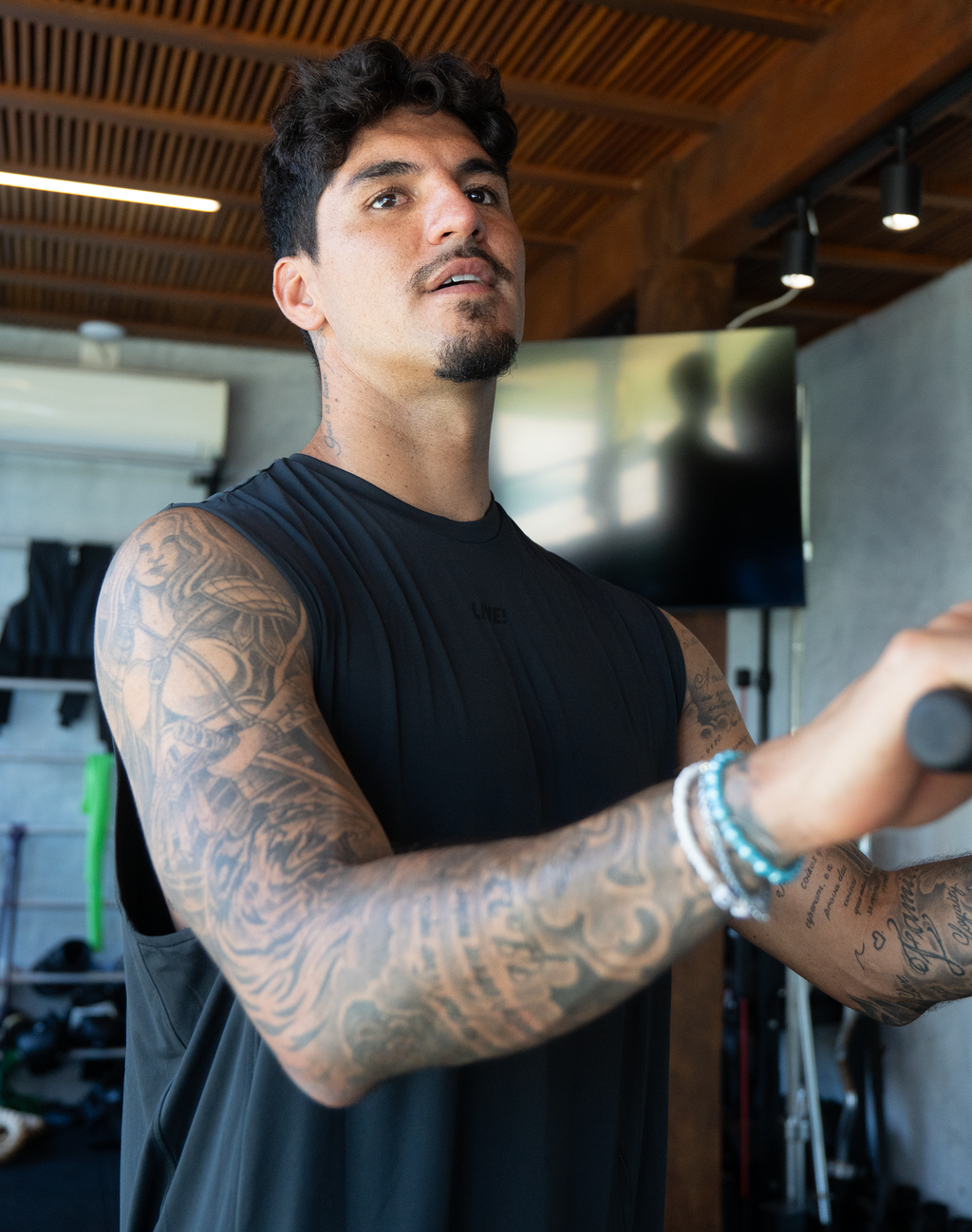 A tattooed man wearing a black sleeveless athletic shirt in a gym setting, holding a barbell.