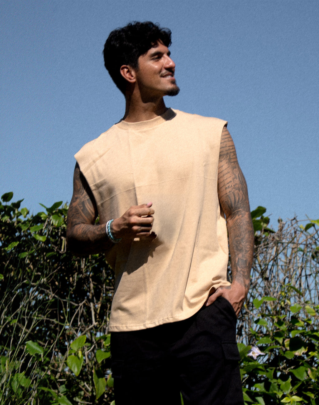 A man wearing a sleeveless beige linen t-shirt and black pants, standing outdoors against a blue sky and green foliage.