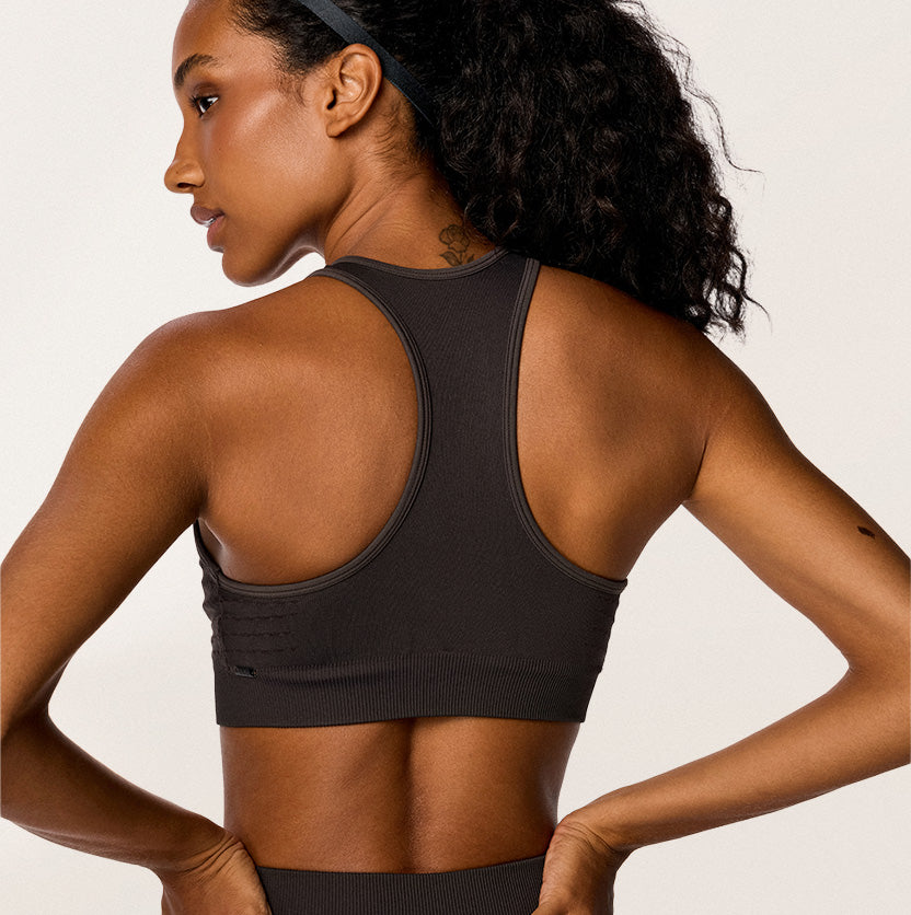 A woman shown from the back wearing a dark charcoal gray athletic sports bra, photographed against a clean off-white studio background.