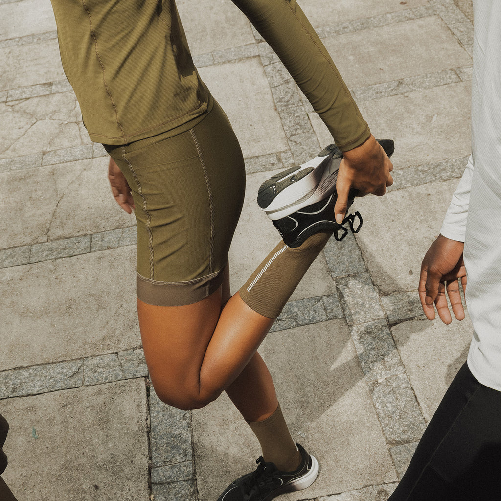 A person in an olive green athletic set stretches their leg while wearing black and white running shoes, standing on a gray stone pavement. Another person is partially visible on the right.