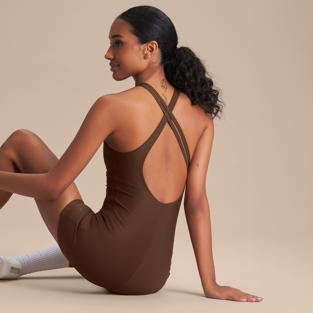 A woman wearing a brown one-piece athletic bodysuit with a crisscross open-back design, seated in profile against a solid beige studio background.