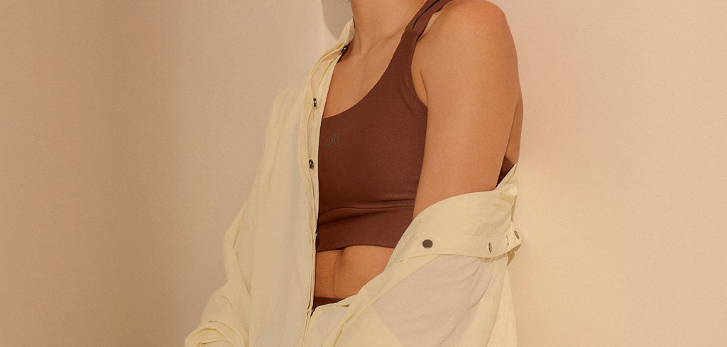 A model wearing a brown ribbed tank top under an open cream-colored button-down shirt, set against a plain beige wall.
