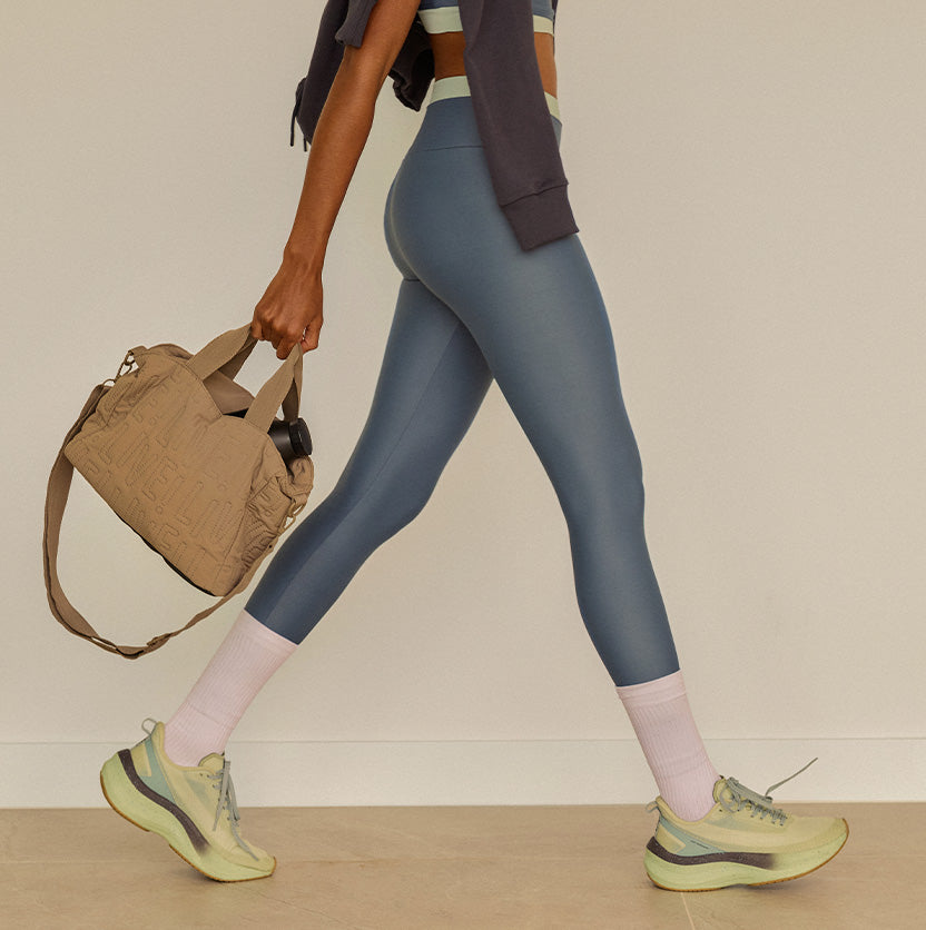 A person walking in athletic wear, shown from the waist down, wearing slate blue leggings, light pink socks, and light green running shoes, while carrying a tan duffel bag against a plain beige wall.