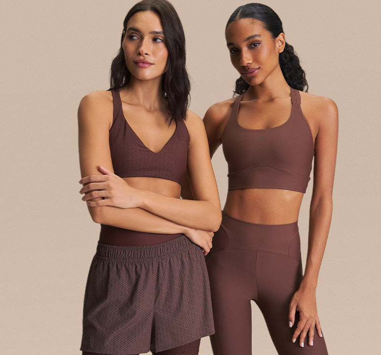 Two women modeling matching athletic wear in a studio setting. The left model wears a textured brown sports bra and shorts set, and the right model wears a solid brown sports bra and leggings set.