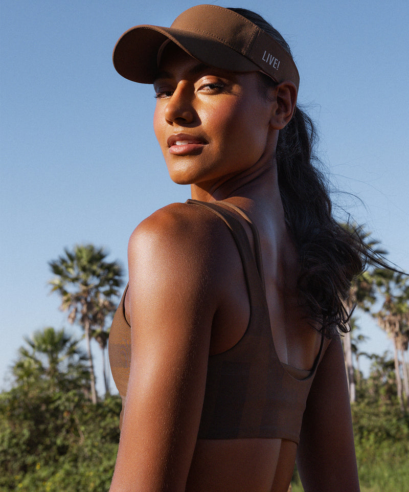 Woman outdoors wearing a brown sports bra and matching visor from LIVE!, with palm trees and blue sky in the background.
