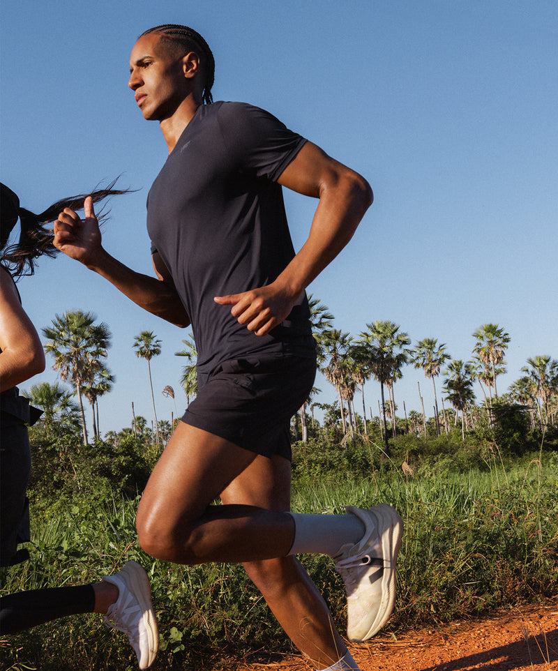 Man running outdoors in a black tee and shorts, wearing white Veja x LIVE! sneakers, surrounded by palm trees and natural vegetation.